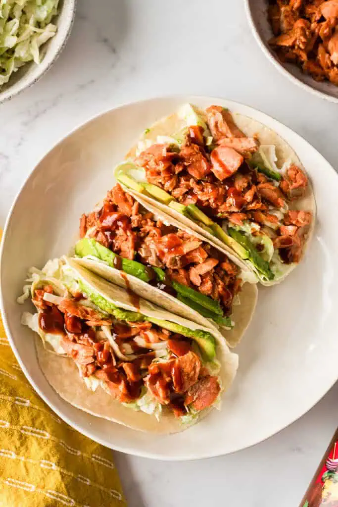 Three barbecue salmon tacos on a plate, a bowl of slaw and bbq salmon in the background.