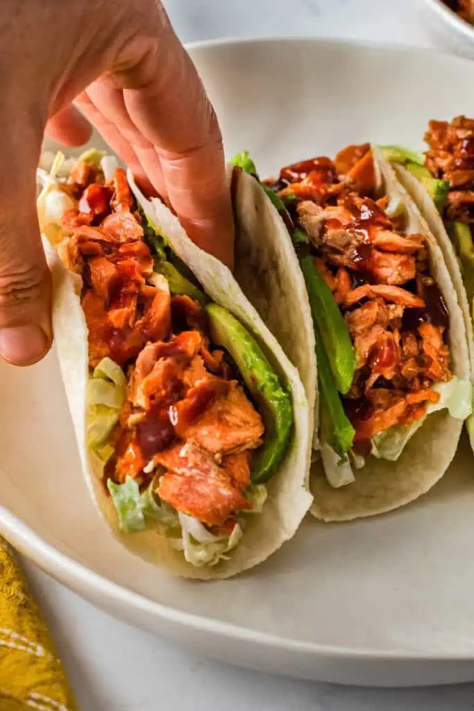 Hand picking up a bbq salmon taco from a white plate.