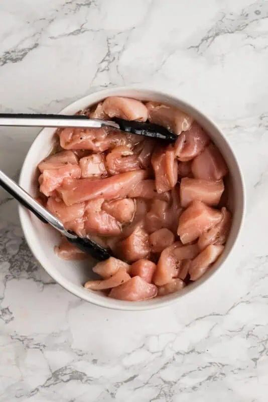 White bowl filled with raw chicken, cubed with dressing on top.