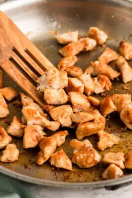 Greek chicken cubes in a skillet, a wood spatula moving them around the pan.