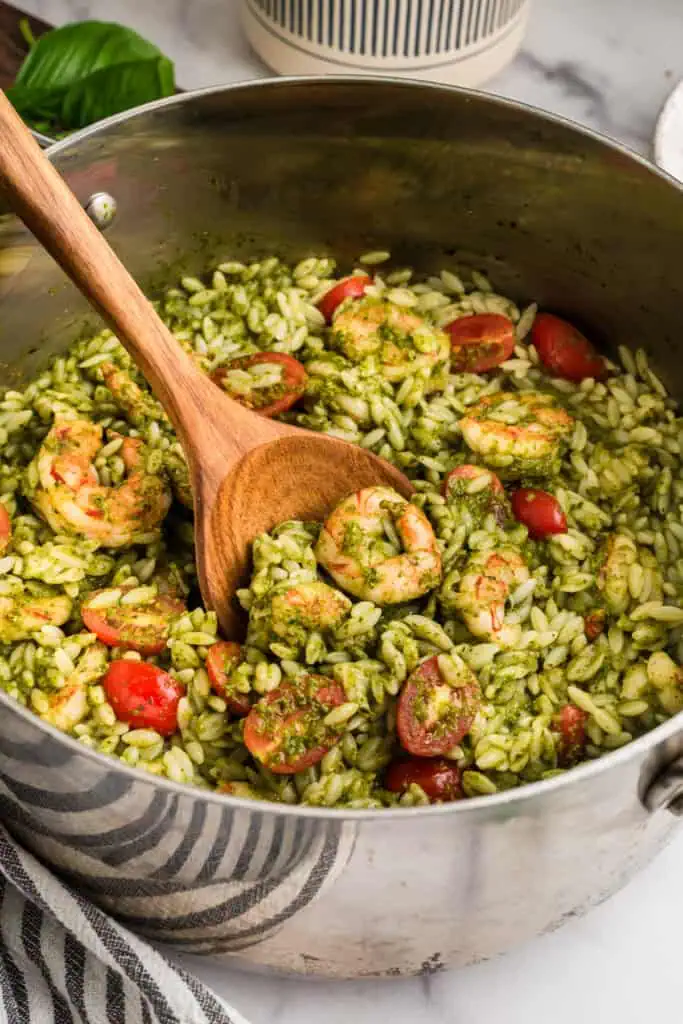 Wood spoon resting in a large stainless steel pot of shrimp orzo pesto.