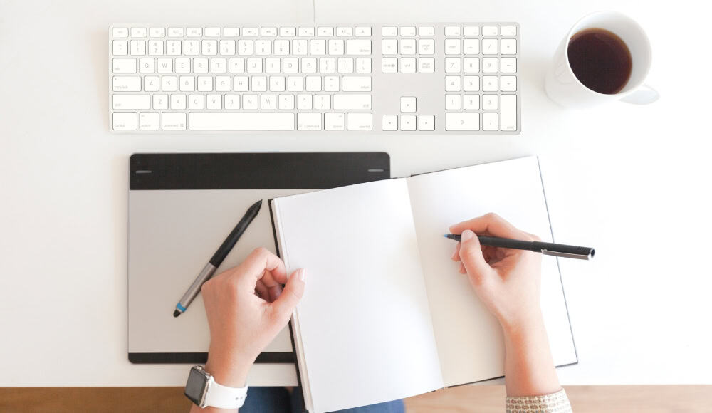 Hands writing in journal near computer keyboard.