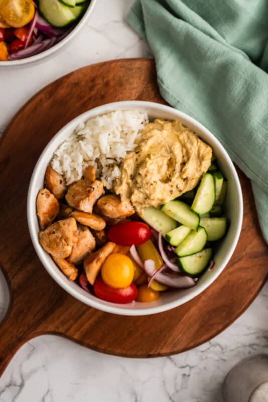 Mediterranean chicken and rice in a bowl with fresh veggies and hummus. Bowl is sitting on a circular wood board.