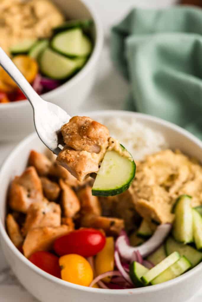 Forkful of Greek chicken and a cucumber being lifted from a bowl.