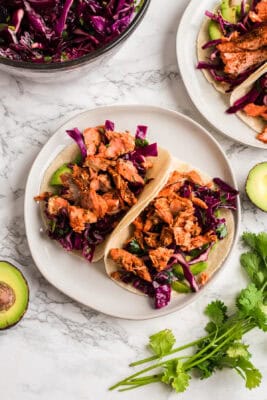 Two chipotle salmon tacos on a white plate with avocado on the side.