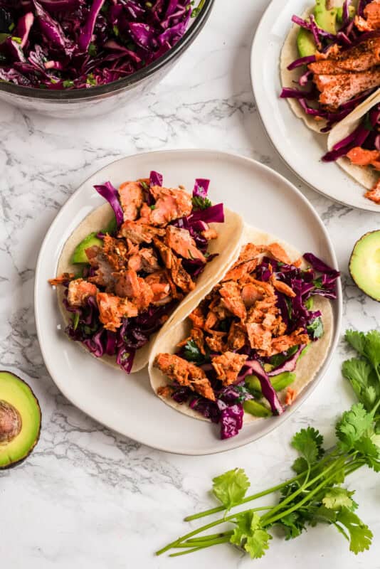 Two chipotle salmon tacos on a white plate with avocado on the side.