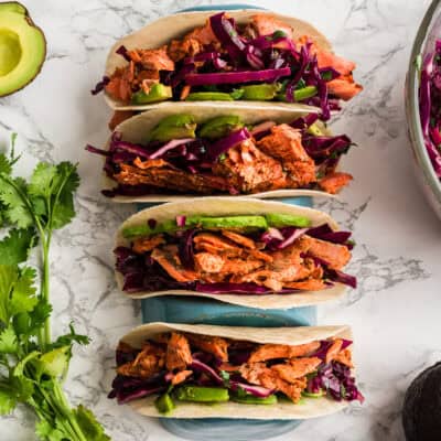 Four chipotle salmon tacos in a blue taco holder on a marble countertop.
