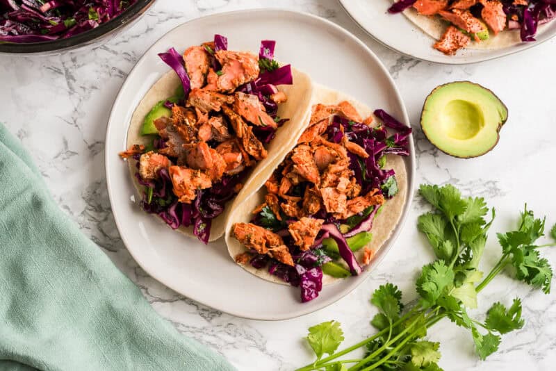 Two chipotle salmon tacos on a white plate with avocado on the side.