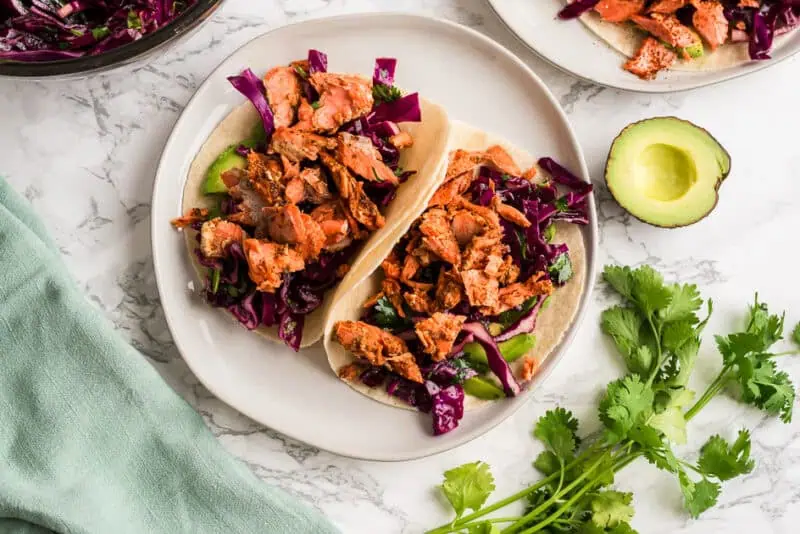 Two chipotle salmon tacos on a white plate with avocado on the side.