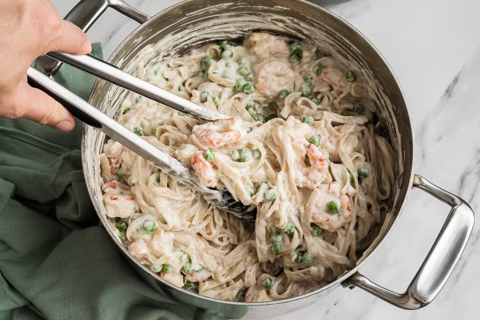 Tongs picking up cottage cheese shrimp alfredo from a pot. 