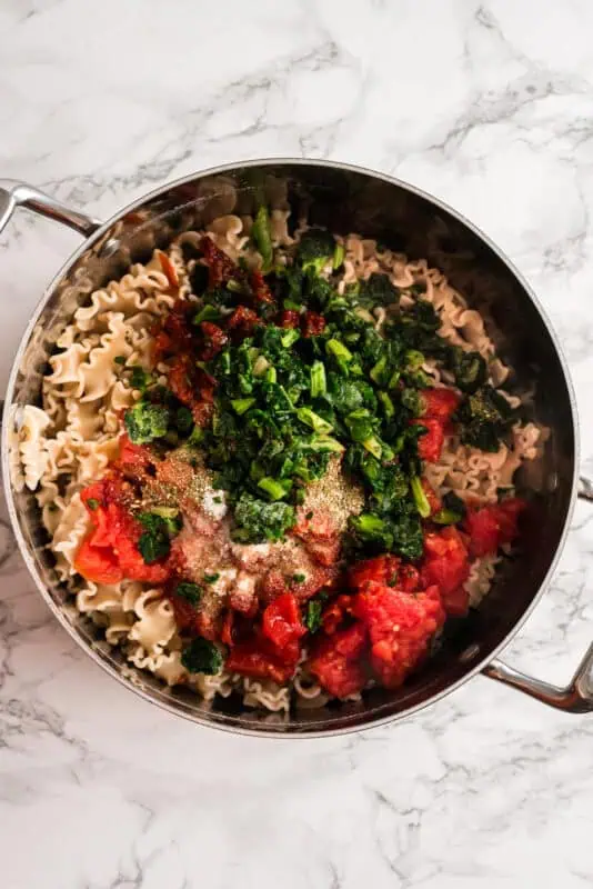 Stainless steel pot filled with pasta, diced tomatoes, frozen spinach, sundried tomatoes, salt and garlic powder.