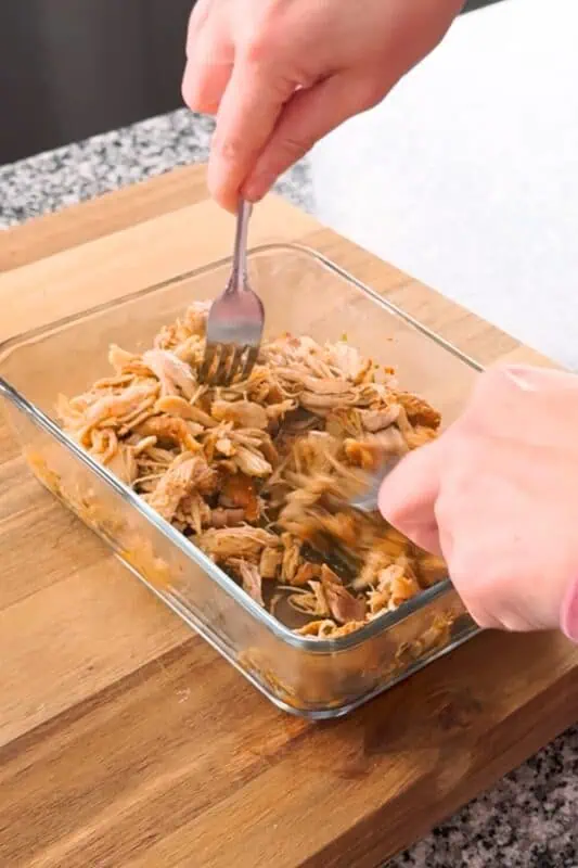 hands holding forks that are shredding chicken in glass container.