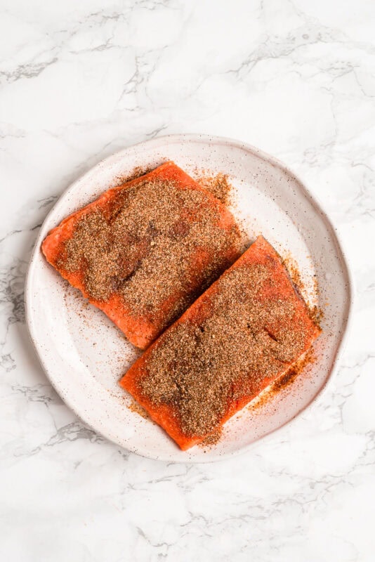 Two salmon fillets with blackening seasoning on them on a white plate.