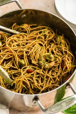 Tongs resting in a large pot filled with pesto shrimp pasta.