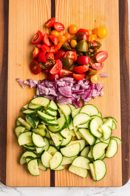Sliced cucumber, chopped red onion and sliced tomatoes on wood cutting board.
