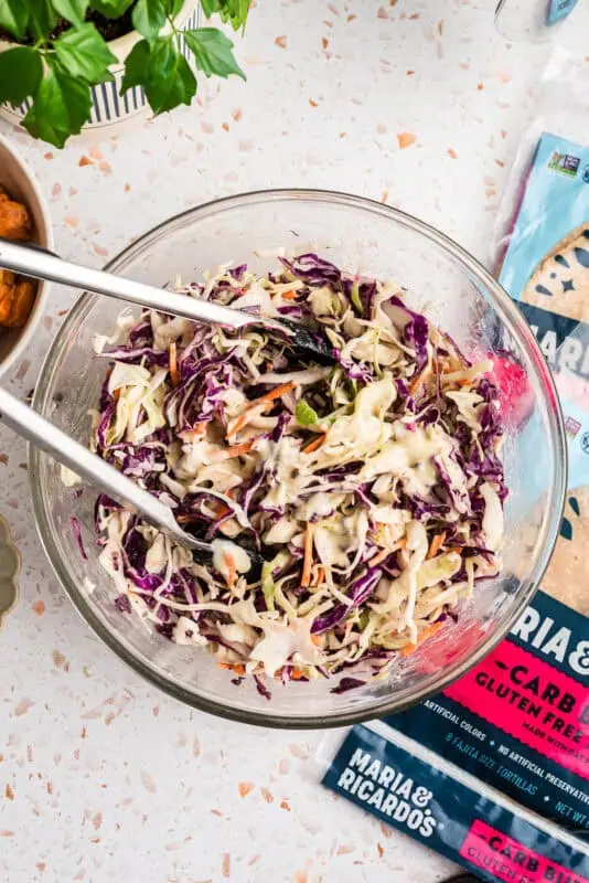 Coleslaw with ranch dressing being tossed in a glass bowl.